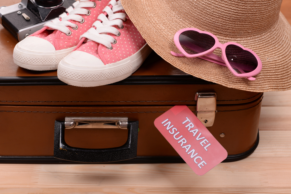 A brown suitcase with a Travel Insurance tag, topped with pink spotty trainers, a straw hat, and pink sunglasses. A camera is partially visible in the background.