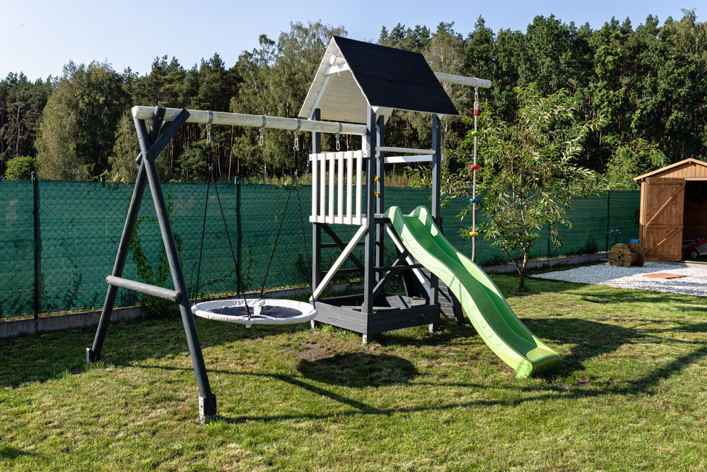 A back garden playset with a small playhouse, slide, rope ladder, round swing, and standard swing, standing on grass, with trees, a shed, and a green fence in the background.