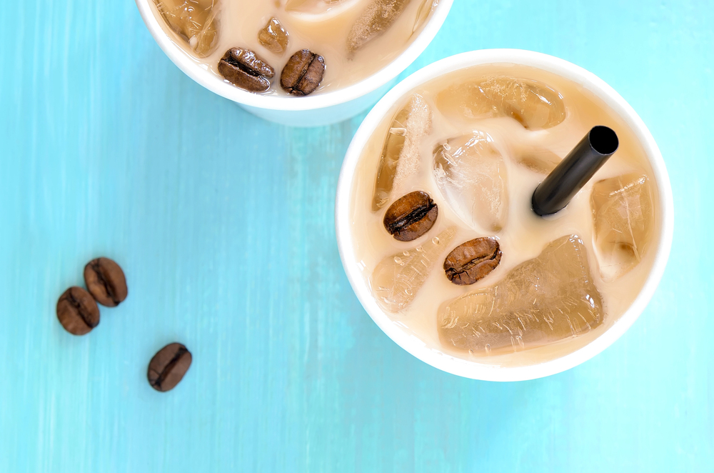Two cups of iced coffee with milk, topped with ice cubes and coffee beans, each with a straw, on a bright blue surface. A few coffee beans are scattered on the table.