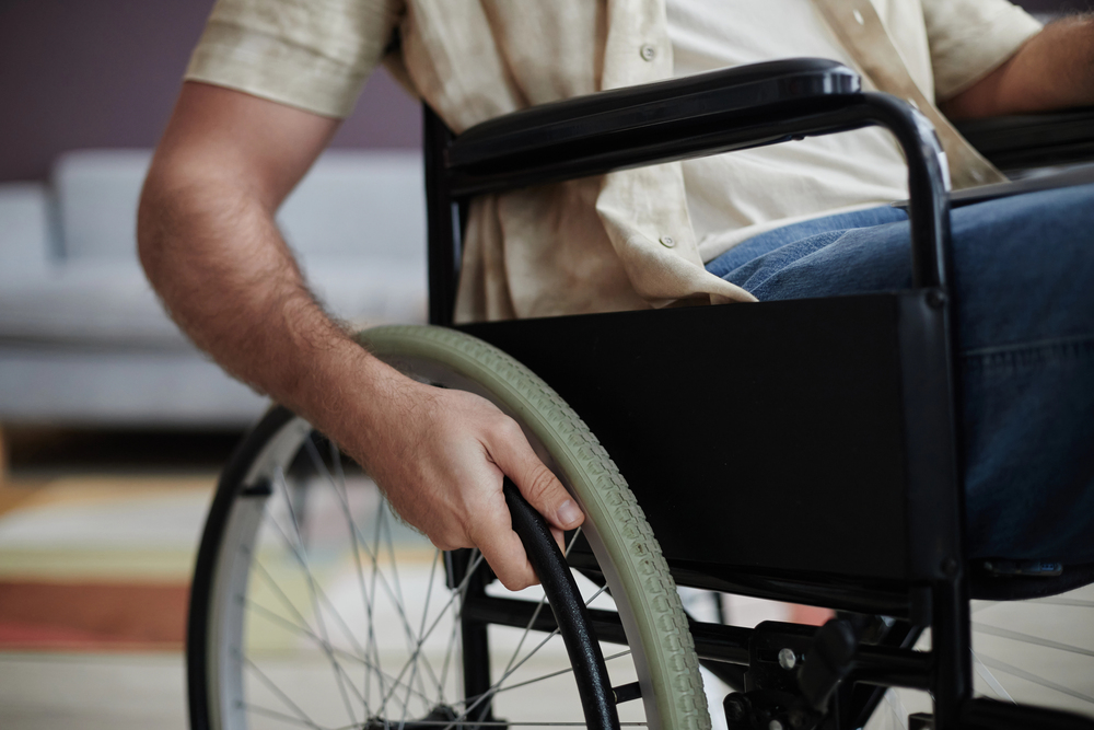 A person in a beige shirt sits in a wheelchair, gripping the wheel with one hand. The background is indoors, with a sofa and soft lighting visible.