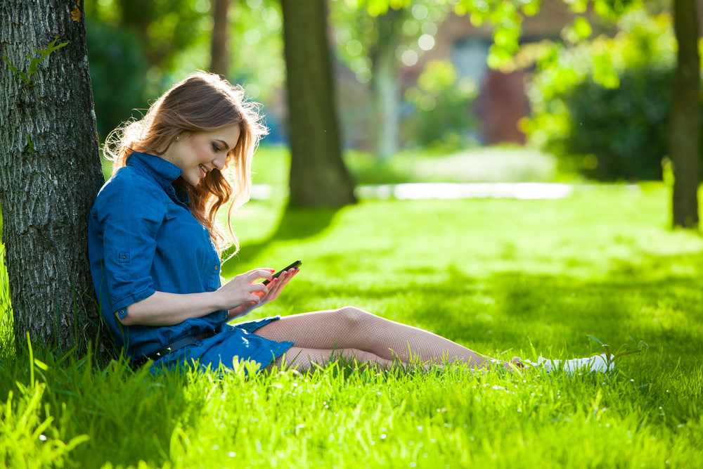 A young woman sits on the grass, leaning against a tree in a sunlit park, smiling whilst looking at her mobile. She is wearing a denim dress and white trainers, surrounded by green grass and trees.