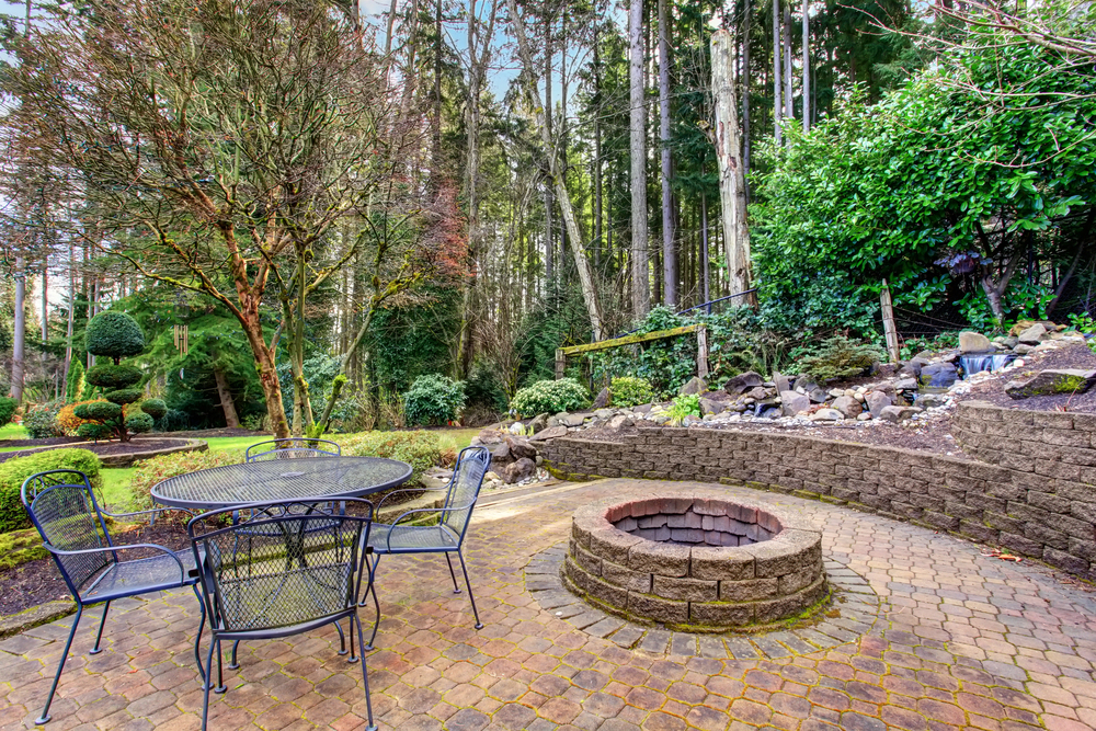 A brick patio with a round fire pit and black metal table with four chairs, surrounded by trees, lush greenery, and a rock garden in a forested back garden setting.