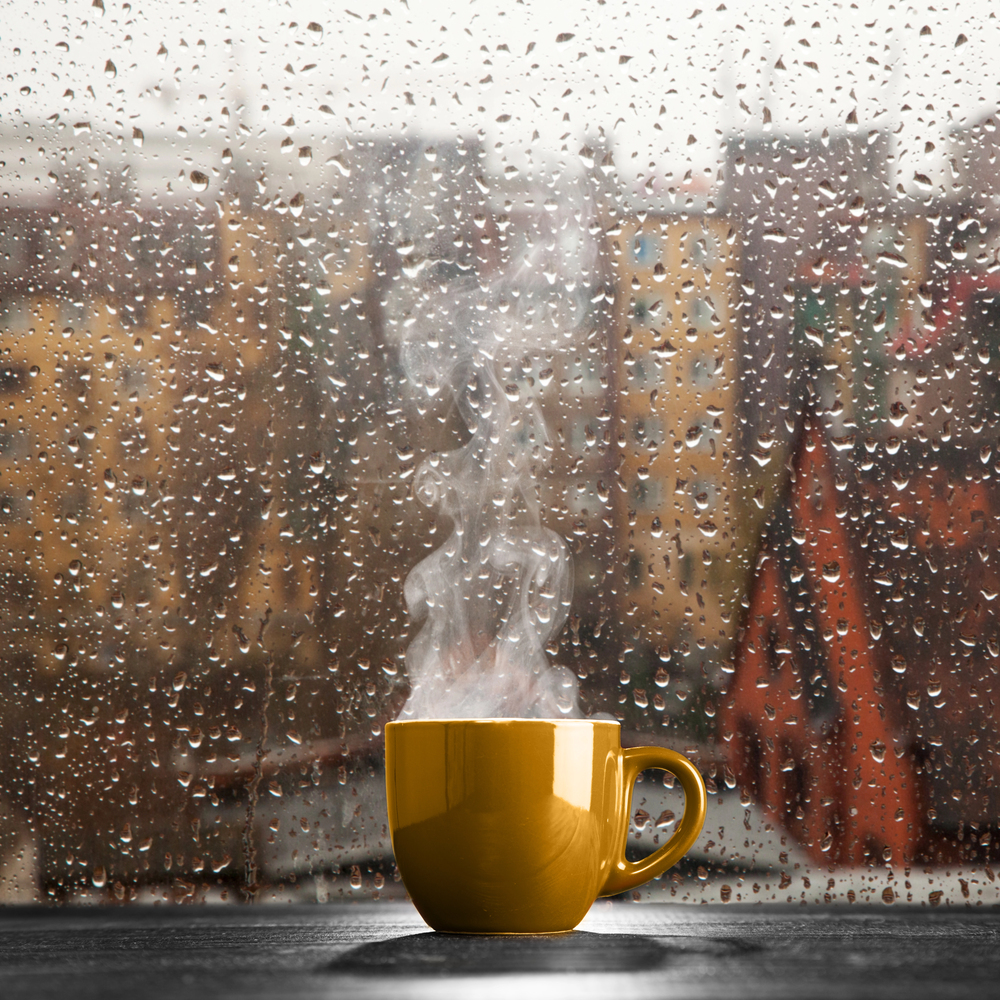 A yellow mug with steam rising from it sits on a windowsill. Outside, raindrops cover the window, and blurred buildings are visible in the background, creating a cosy, rainy day atmosphere.