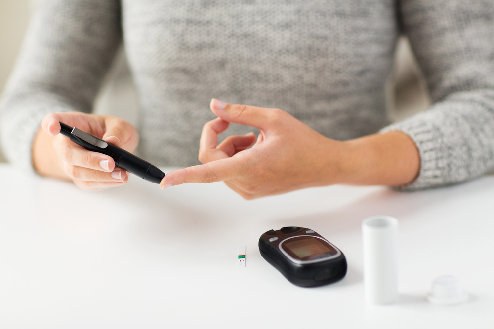 A person pricks their finger with a blood glucose meter whilst sitting at a white table. A test strip, glucose monitor, and a small white container are also on the table. The person is wearing a grey jumper.
