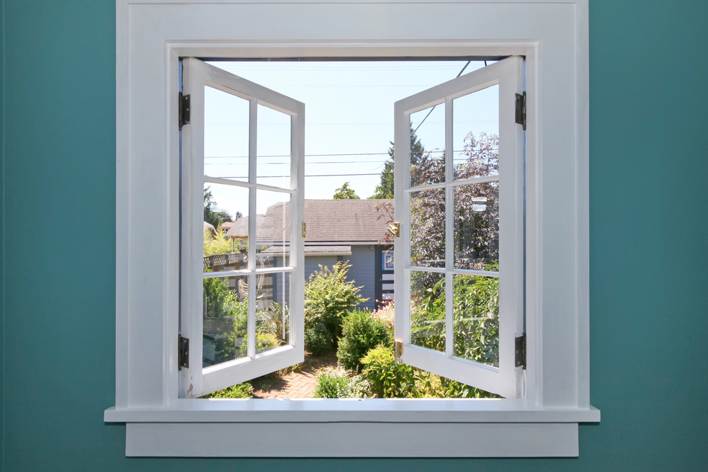 An open white-framed window set in a teal wall looks out onto a sunny garden with green plants, shrubs, and a blue house in the background, under a clear sky with visible power lines.