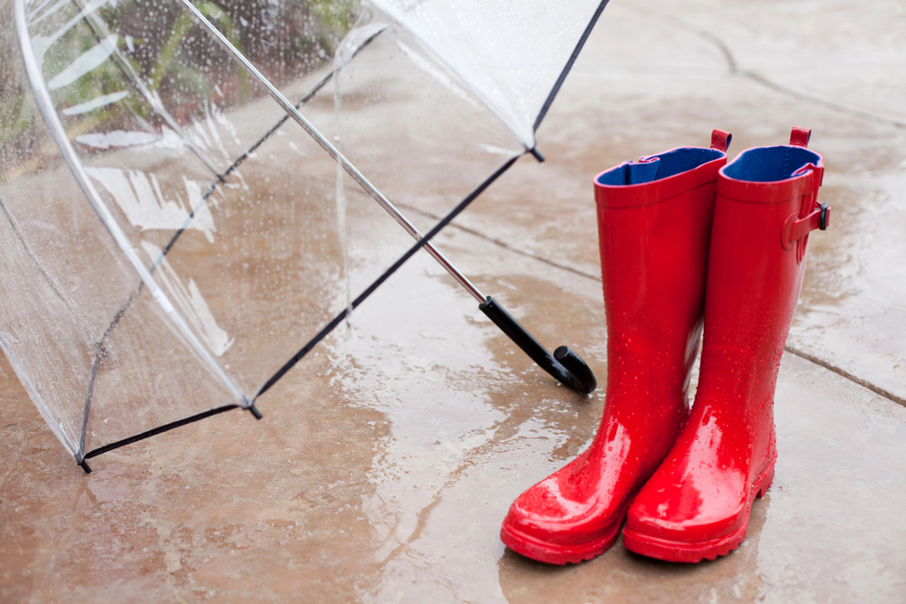 A pair of bright red wellies and a clear umbrella rest on a wet pavement, with raindrops visible on the wellies and ground.