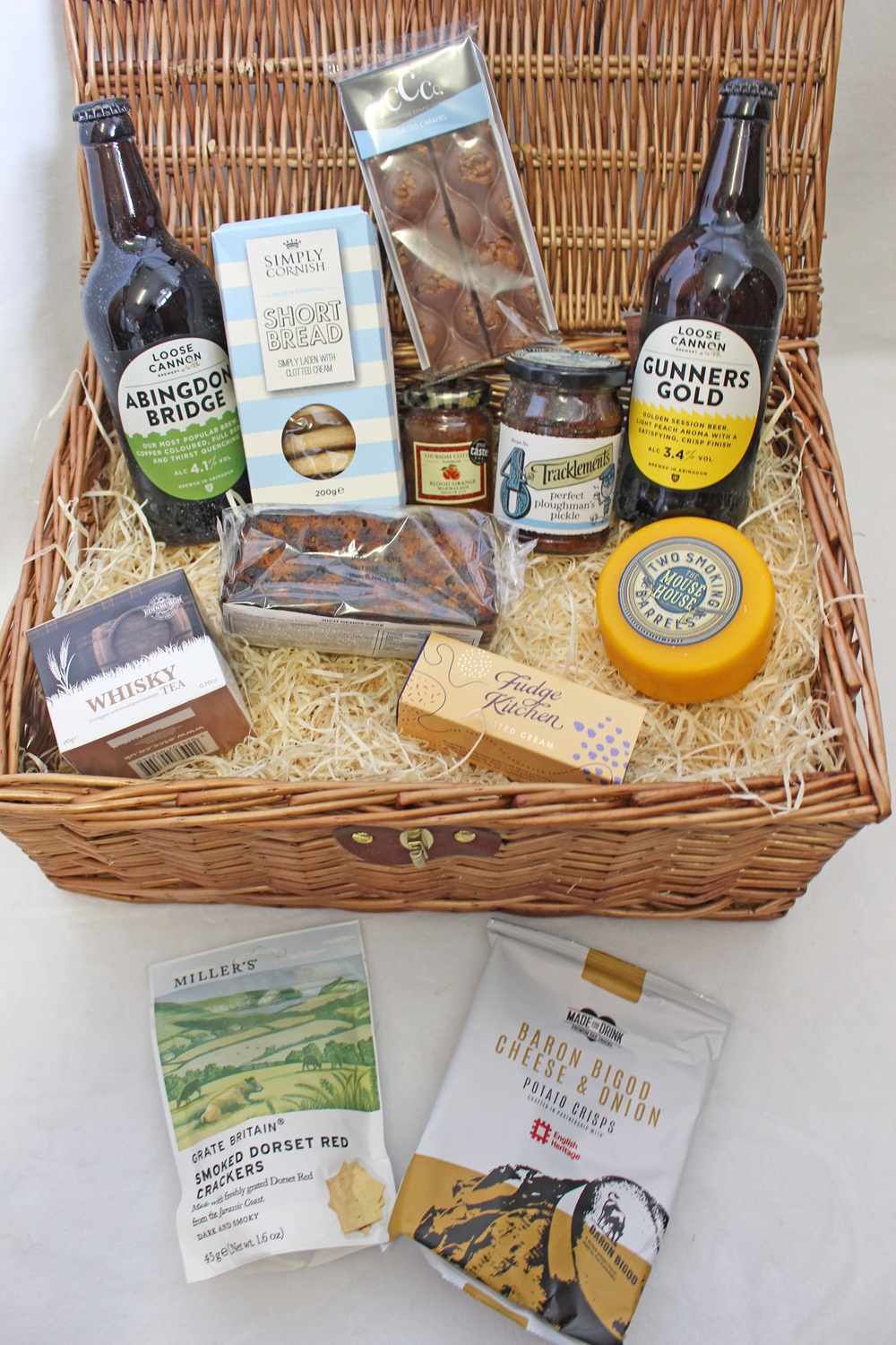 A wicker hamper filled with assorted gourmet items, including bottles of cider, shortbread, biscuits, cheese, chutney, chocolate, and other snacks, all arranged on straw-like packing material.