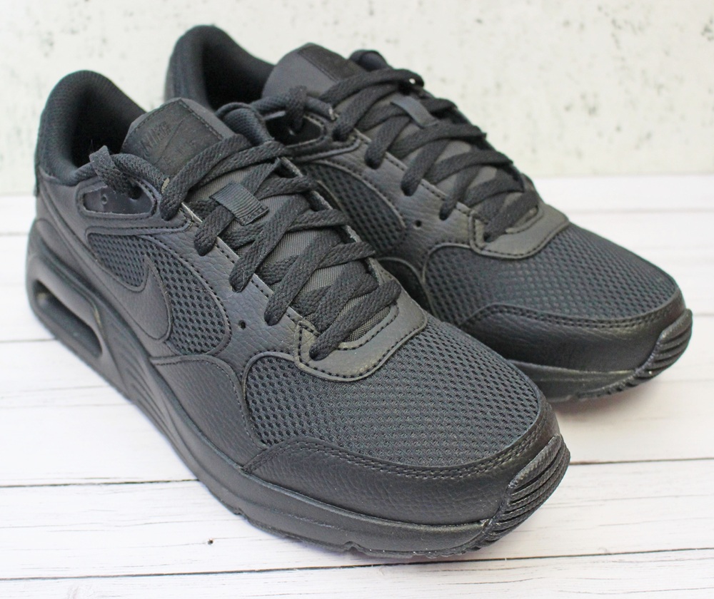 A pair of black athletic trainers with mesh and leather details, black laces, and thick rubber soles, placed on a light wooden surface.