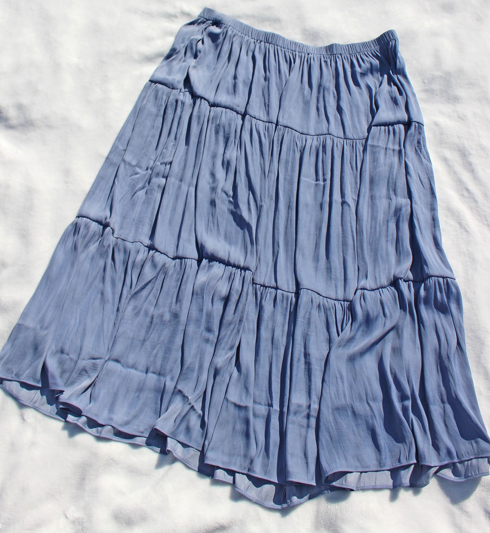 A light blue, tiered midi skirt with a flowing, crinkled texture is laid flat on a white surface. The skirt has an elasticated waistband and features three gathered tiers for added volume.