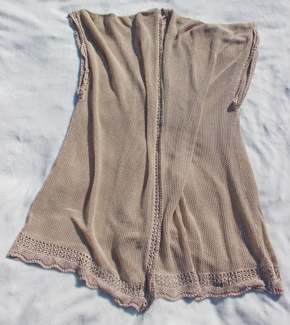 A light beige, sleeveless knitted waistcoat with lace trim along the hem and edges is laid flat on a white surface. The fabric appears soft and slightly textured.