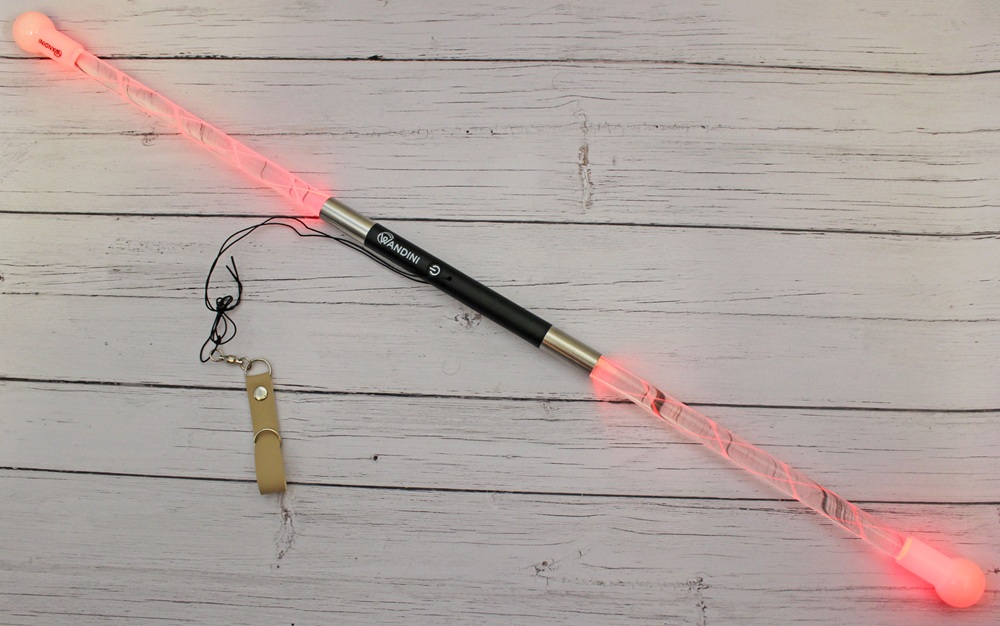 A light-up baton with glowing red ends and a black handle, lying on a light wooden surface next to a beige whistle on a lanyard.