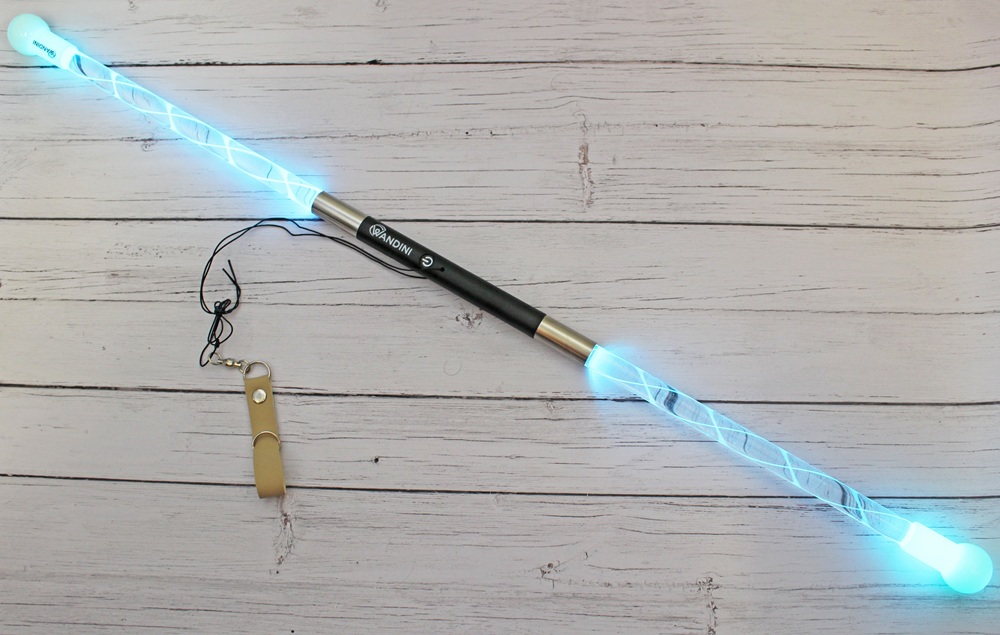 A light-up flow staff with glowing blue ends and a spiral design lies on a wooden surface. A black strap and a tan case or cover are placed nearby.