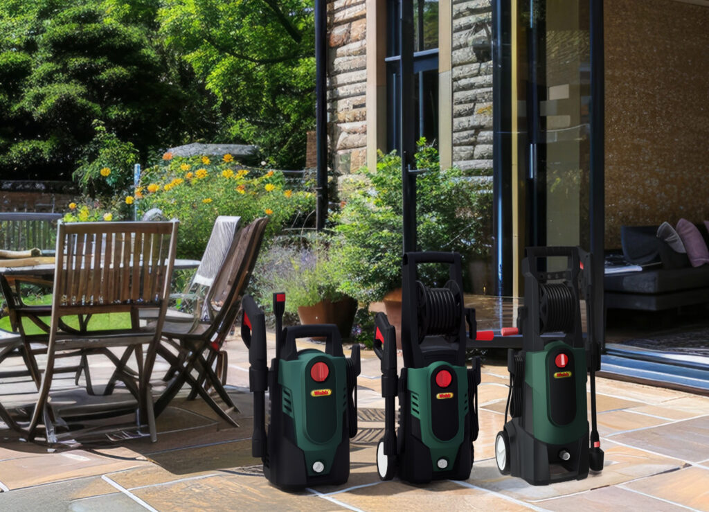 Three green and black pressure washers are placed on a sunny stone patio near wooden garden furniture, flowers, and an open glass door leading to a modern living area.