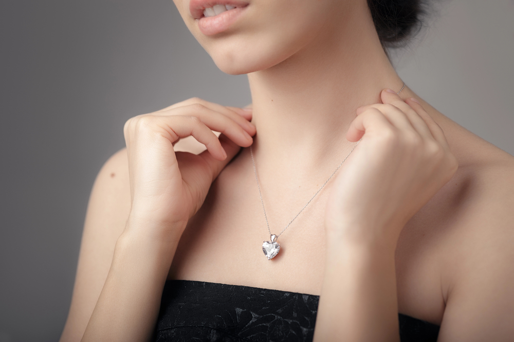 A woman wearing a strapless black top gently holds a silver chain necklace with a heart-shaped pendant around her neck. The background is plain and neutral.
