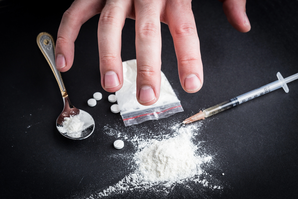 A hand reaches towards a pile of white powder, tablets, a small plastic bag of powder, a spoon with residue, and a syringe, all on a dark surface, suggesting drug use.