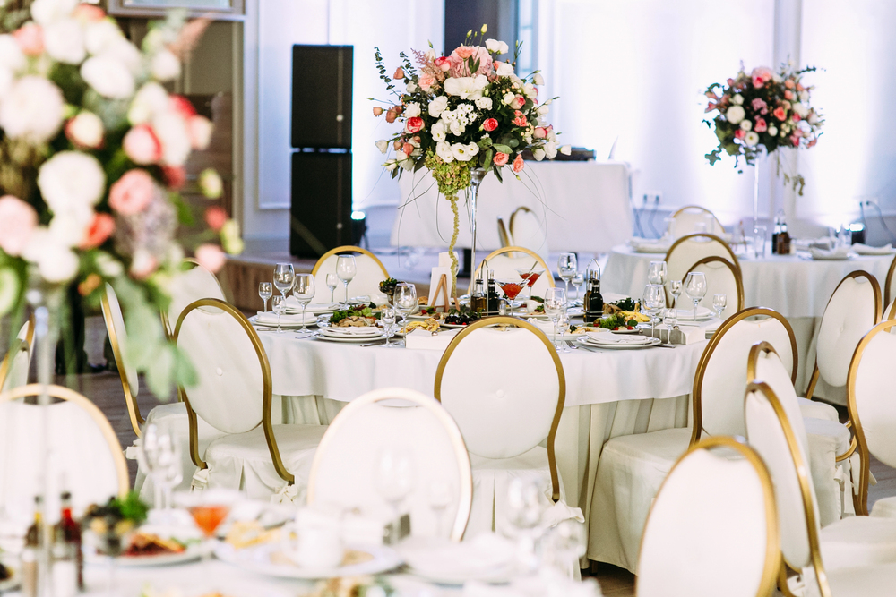 Elegant banqueting hall with round tables covered in white tablecloths, gold-rimmed chairs, and tall floral centrepieces featuring pink and white flowers; tables are set with glassware, plates, and food.