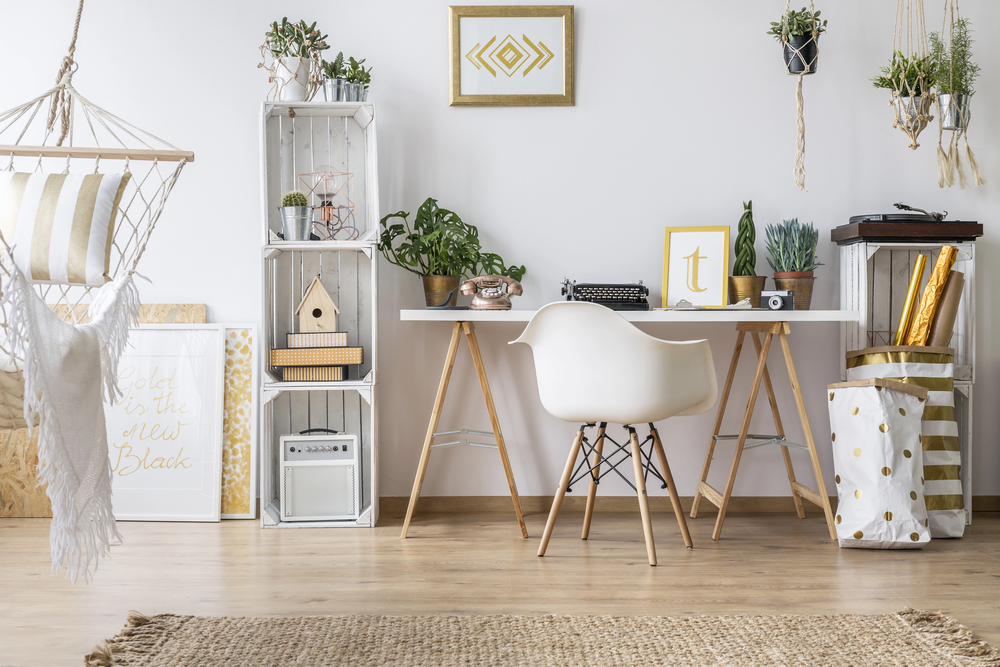 A modern, cosy home office with a white desk, white chair, potted plants, decorative shelves, a typewriter, camera, framed art, and gold-accented decor. Natural light and wooden flooring create a warm atmosphere.