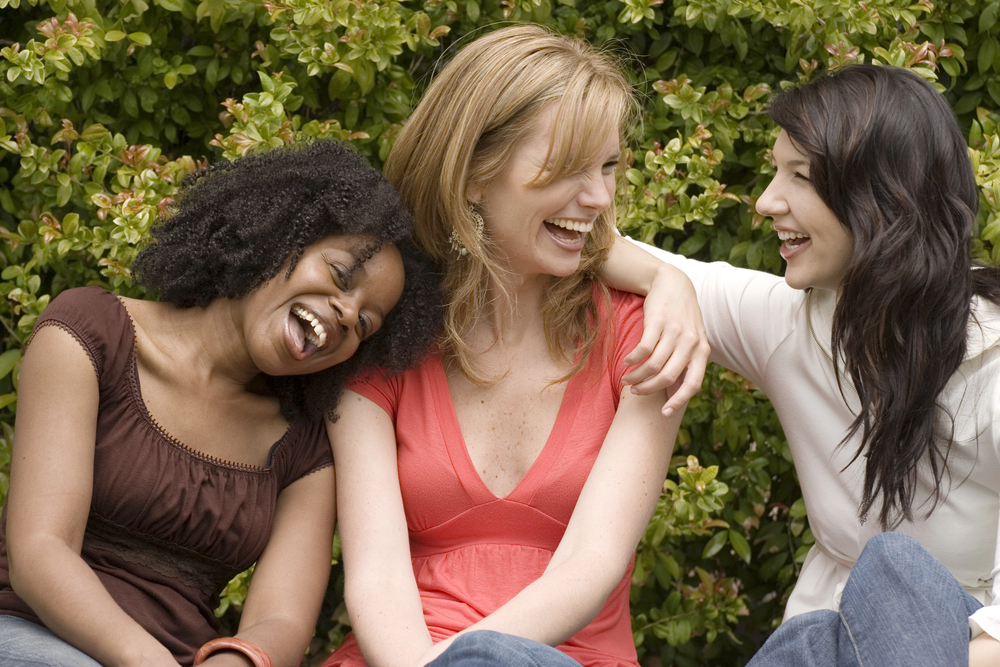 Three women sitting closely together outdoors, laughing and smiling, with one woman resting her arm around the others’ shoulders. Green bushes fill the background, creating a friendly and joyful atmosphere.