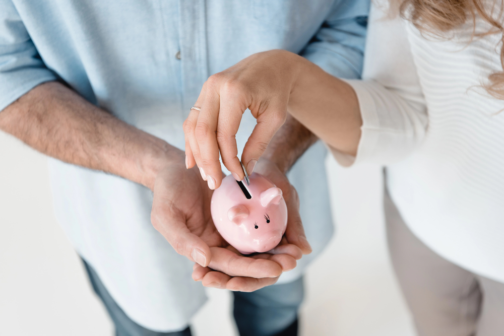 A close-up view of two people from above; one holds a pink piggy bank in their hands whilst the other drops a coin into it, symbolising saving money together.