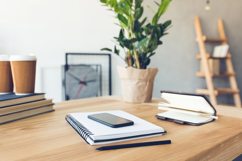 A tidy wooden desk with a spiral notebook, mobile phone, pencil, tablet cover, and takeaway coffee cups. A potted plant, stack of books, wall clock, and wooden ladder shelf are in the background.