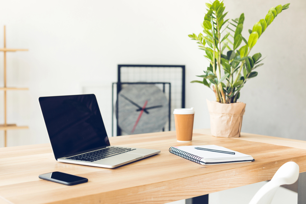 A modern workspace with a laptop, mobile phone, notebook, and pen on a wooden desk, a takeaway coffee cup, and a potted plant. A decorative clock leans against the wall in the background.