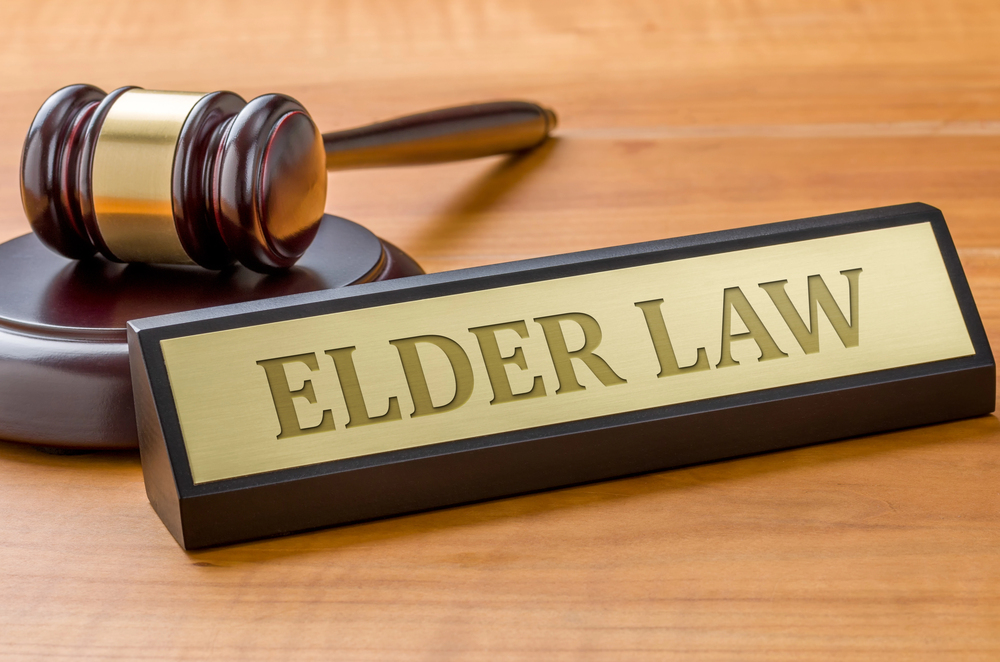 A wooden judge’s gavel rests beside a desk nameplate that reads “ELDER LAW” in bold letters on a gold background, all placed on a wooden surface.