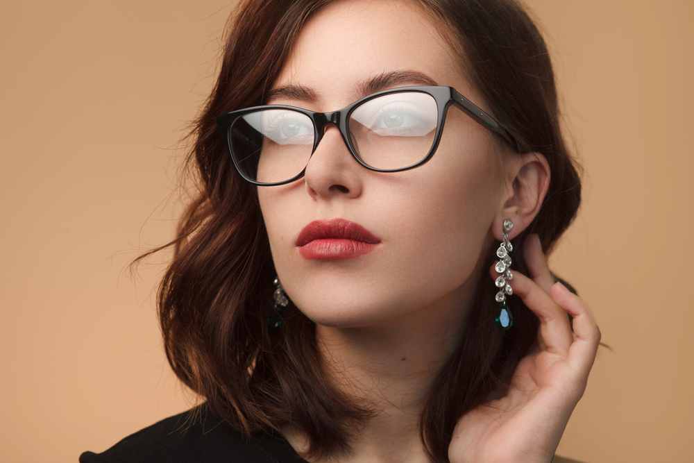 A woman with shoulder-length brown hair, red lipstick, and dangling earrings wears black glasses steamed up over the lenses. She holds one hand near her ear against a beige background.