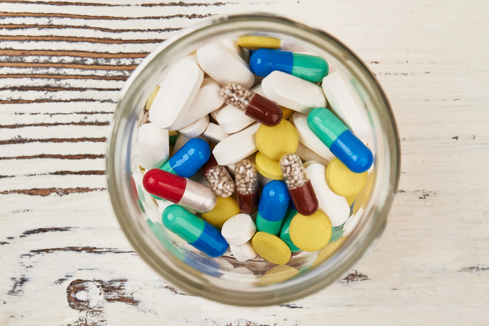 A glass jar filled with assorted pills and capsules of various shapes, sizes, and colours, including white, yellow, green, blue, and red, sitting on a rustic wooden surface.