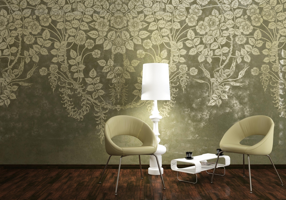 Two modern beige chairs and a small white table with a cup sit on a dark wooden floor next to a tall white lamp. The background features a green wall with an ornate floral pattern.