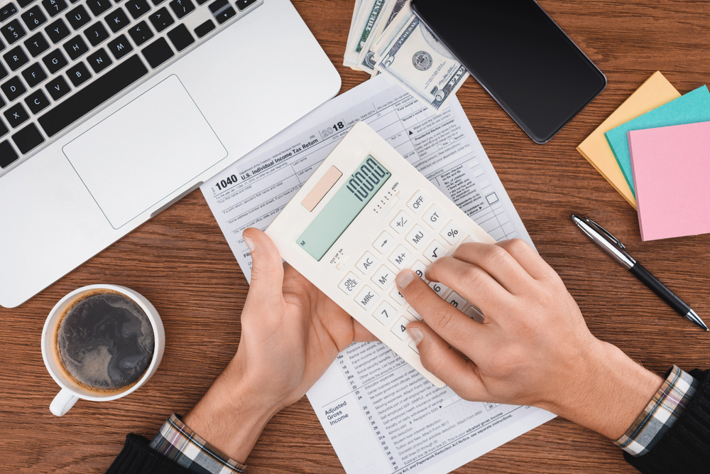 A person uses a calculator on top of tax forms at a desk with a laptop, cash, mobile phone, pen, sticky notes, and a cup of coffee.