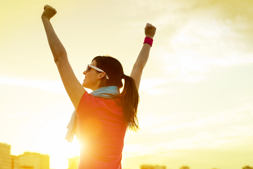 A woman wearing sunglasses raises her arms in triumph at sunrise or sunset, with sunlight shining behind her and city buildings in the background.