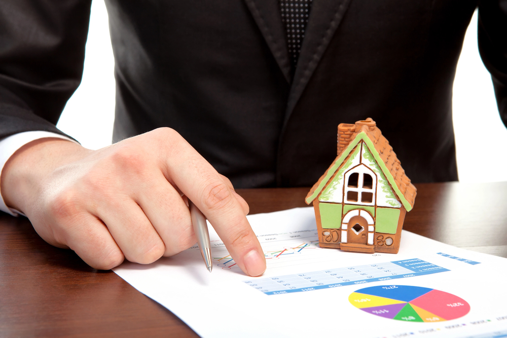 A person in a suit points at a financial chart on paper with a pen, next to a small model house, suggesting property or mortgage analysis.