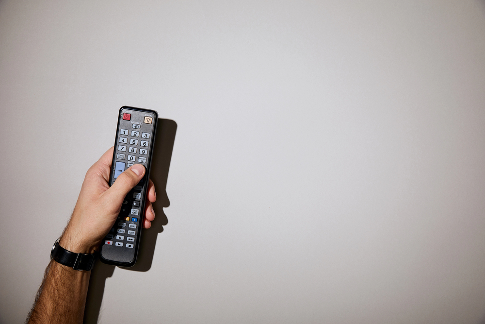 A person wearing a watch holds a black TV remote control, pointing it towards a blank white wall.