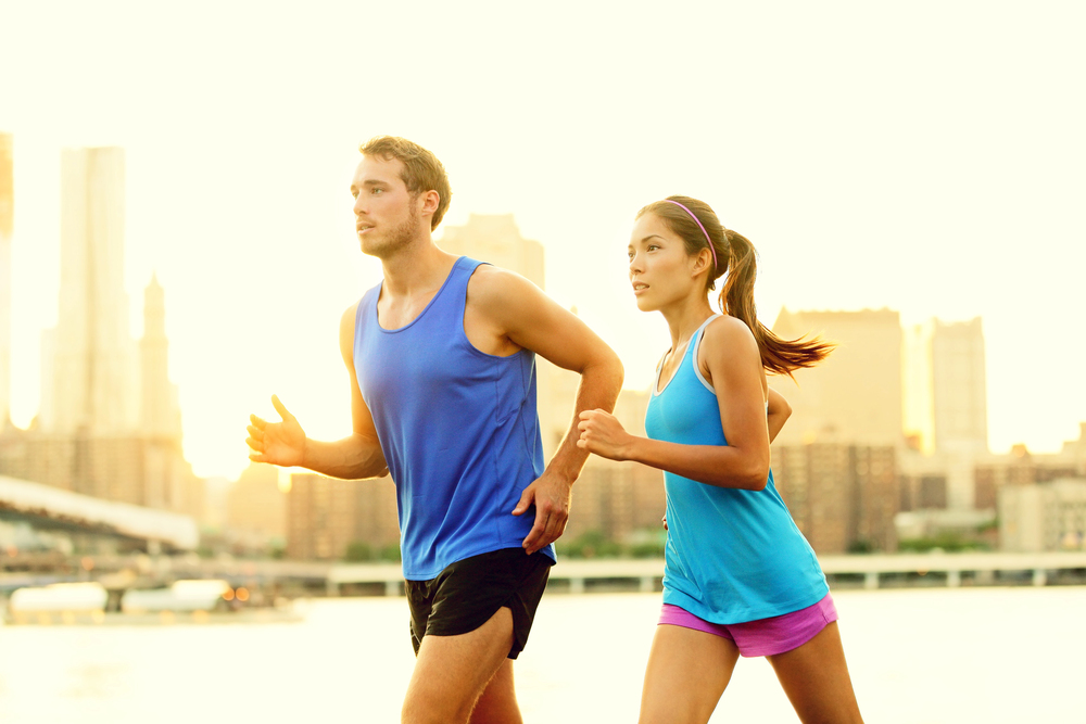 A man and woman in sportswear jogging outdoors by a waterfront, with a city skyline and buildings in the background at sunset.