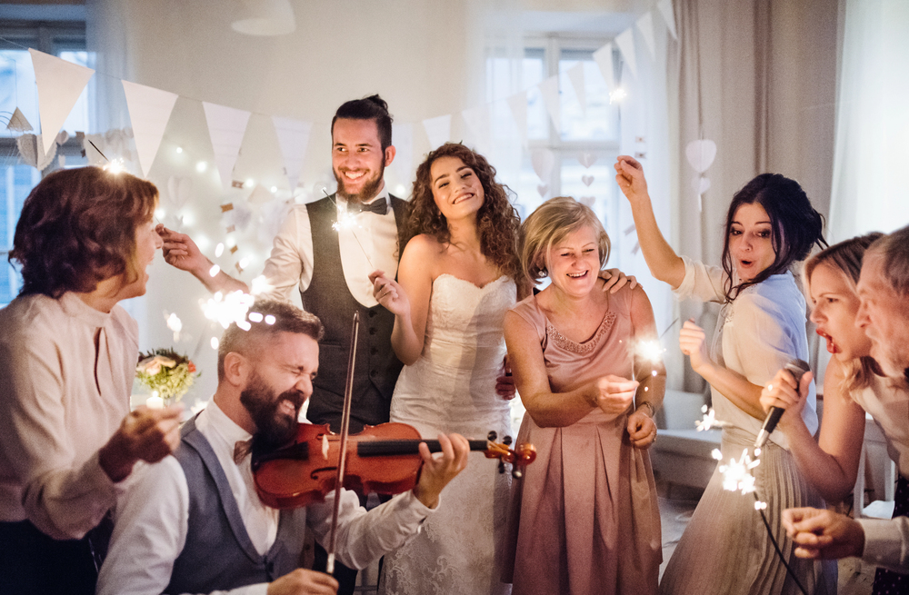 A joyful bride and groom celebrate with friends and family indoors, waving sparklers while a violinist plays. Everyone is smiling and dressed in formal attire, surrounded by festive decorations and white bunting.