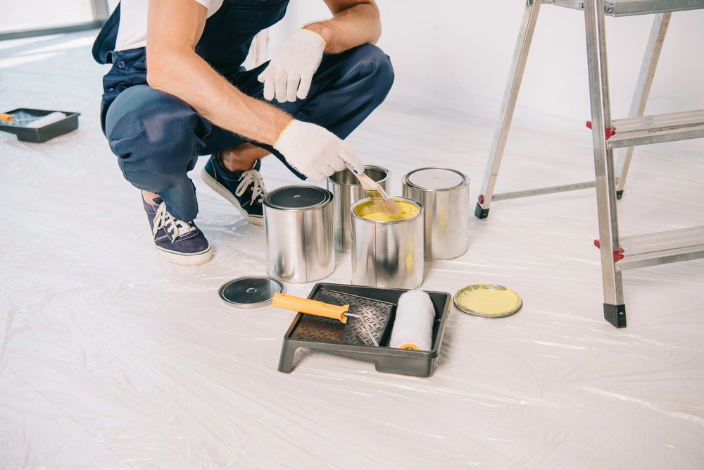 A person wearing gloves and overalls kneels on the floor, stirring yellow paint in a tin. Paint tins, a roller, a tray, and a ladder are nearby. The floor is covered with plastic sheeting for protection.