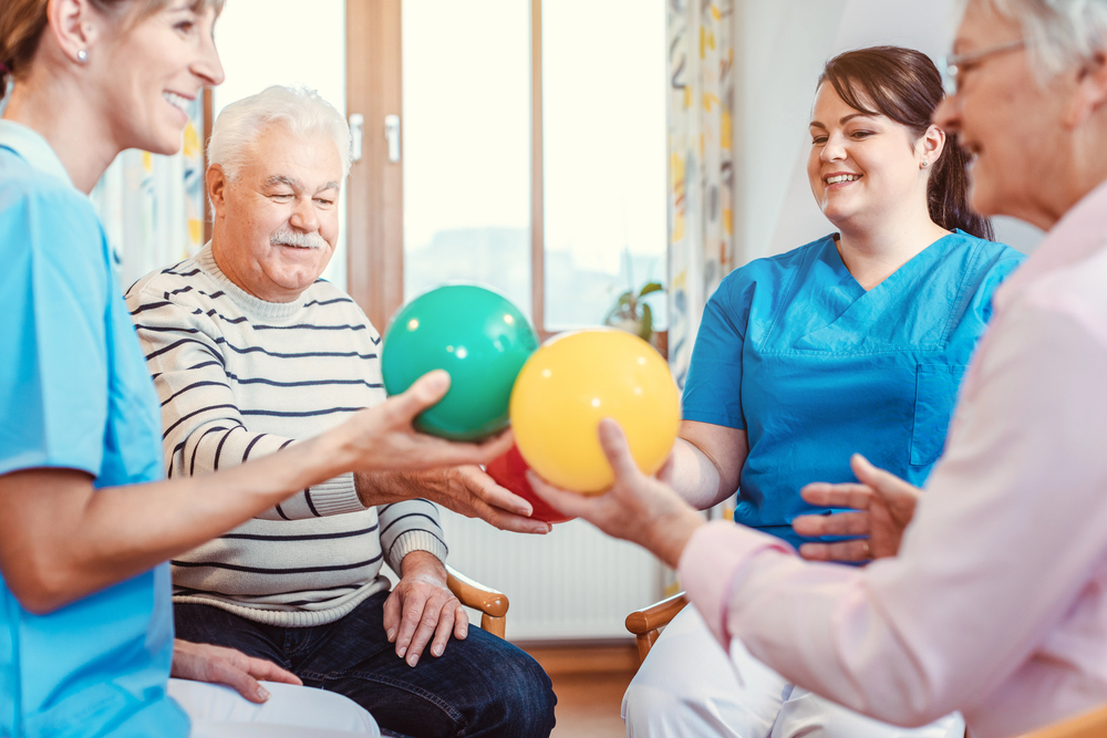 Four people, including two elderly adults and two carers in blue uniforms, sit in a circle indoors, smiling and holding colourful exercise balls, engaging in a group activity.