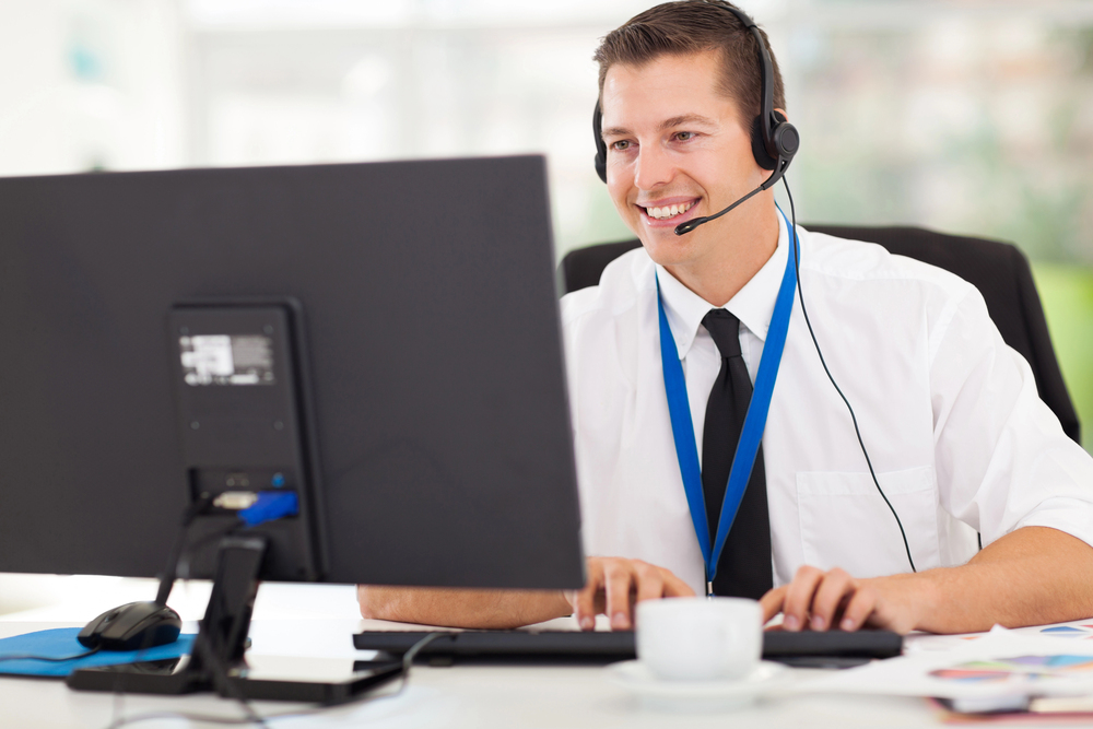 A man wearing a white shirt, tie, and headset with a microphone sits at a desk, smiling whilst working on a computer. A coffee cup is on the desk, and he wears a blue lanyard round his neck.