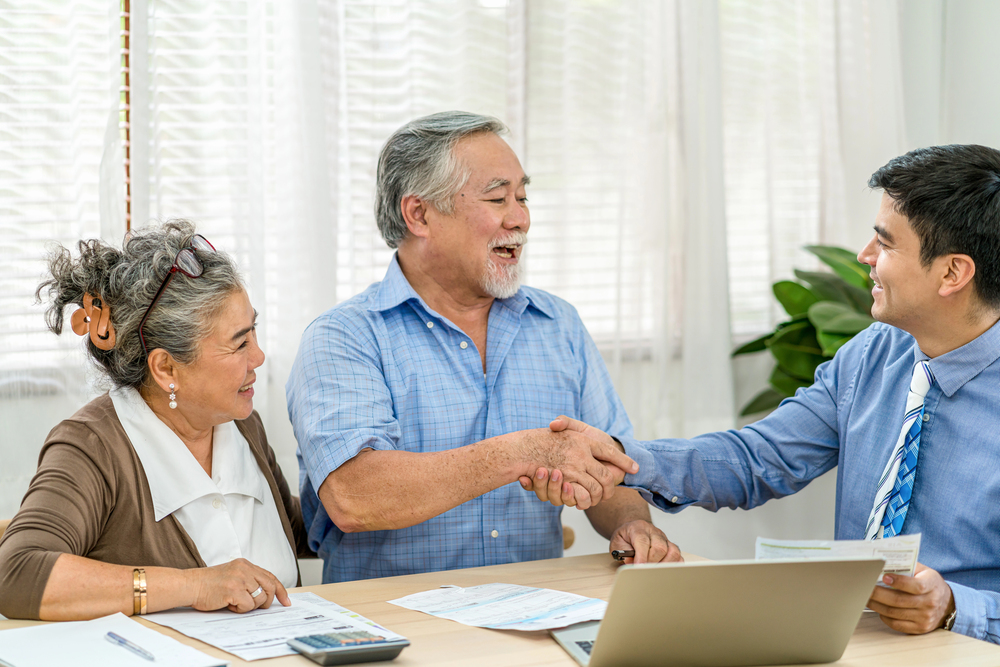 An older couple sits at a desk with documents, smiling as the man shakes hands with a younger man in a suit. A laptop, pen, and calculator are on the table in a bright office.
