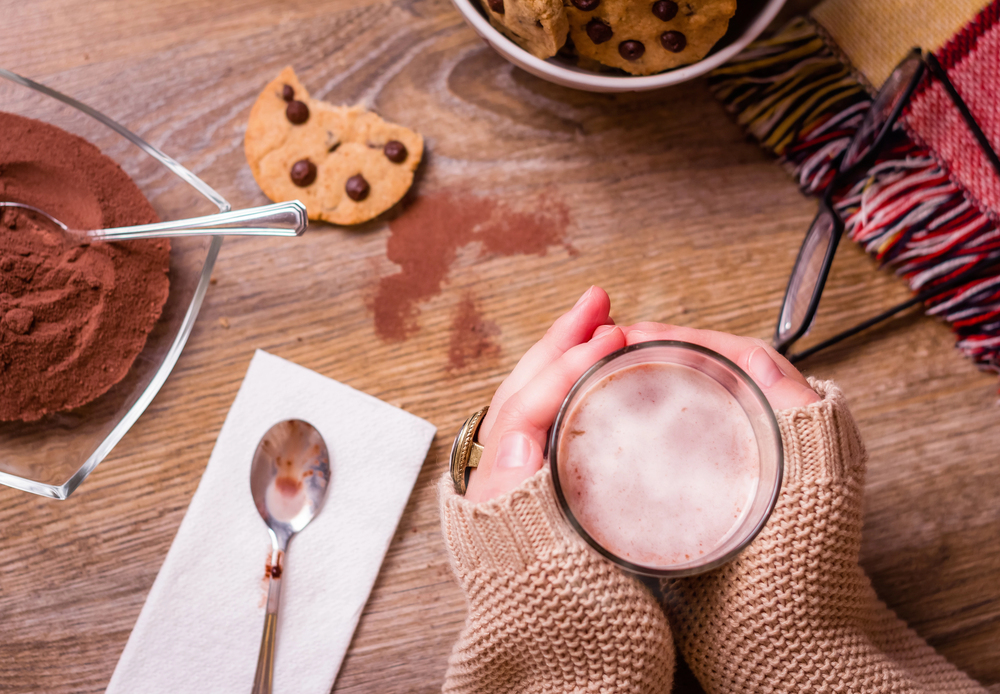 A person in a cosy jumper holds a glass of hot chocolate on a wooden table with chocolate chip biscuits, a bowl of cocoa powder, a spoon on a serviette, and a colourful scarf.
