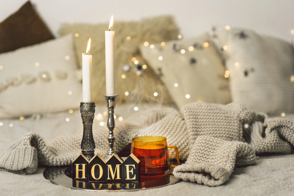 Two lit candles in silver holders and a mug of tea sit on a tray with wooden blocks spelling HOME, surrounded by cosy knitted blankets and cushions, with warm fairy lights in the background.