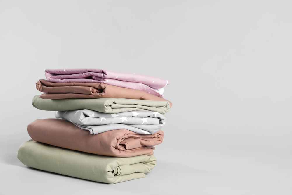 A stack of neatly folded bed sheets in pastel colours, including pink, green, beige, and grey, with some featuring small white star patterns, set against a plain light grey background.