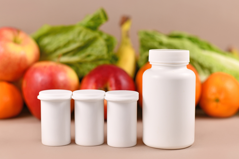 Four white pill bottles of varying sizes are lined up in front of assorted fresh fruit and leafy greens against a neutral background.