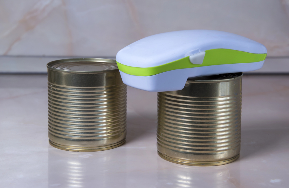 A handheld electric tin opener rests on top of two closed, unlabelled tins on a light-coloured worktop with a marble-patterned background.