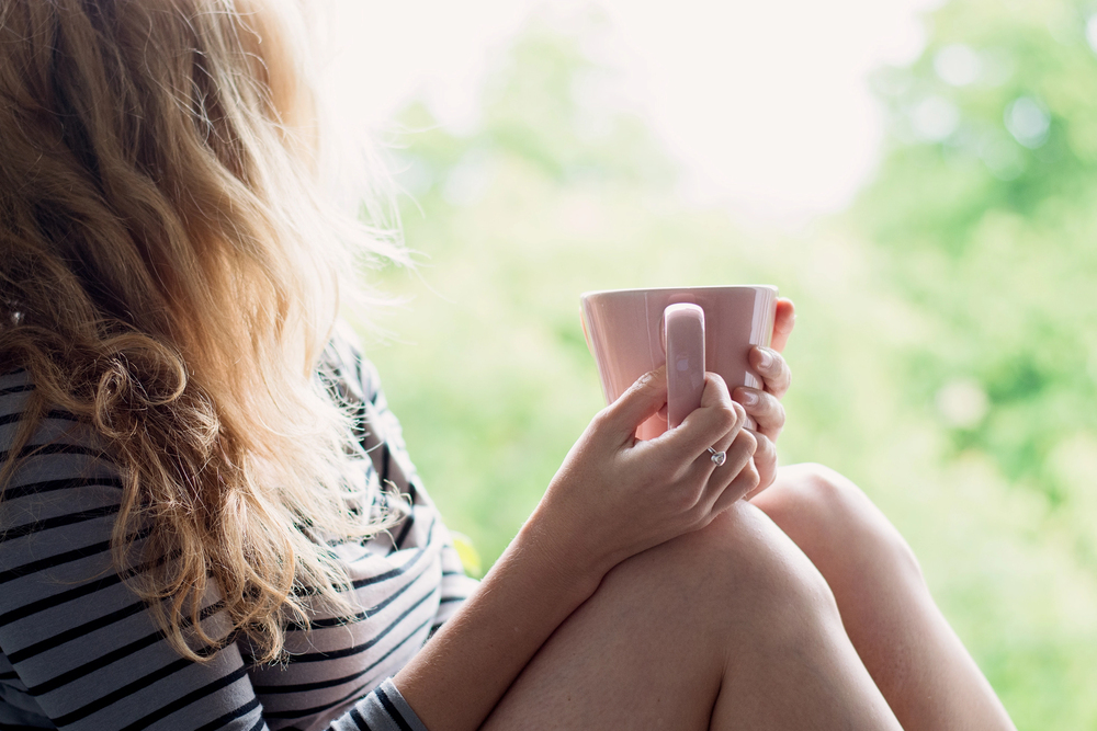 A woman with wavy blonde hair sits by a bright window, holding a pink mug in both hands. She wears a striped long-sleeve shirt and has her knees drawn up, with greenery visible outside.