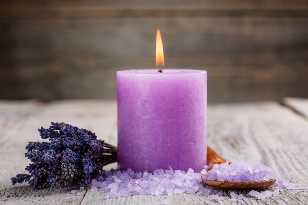 A lit purple candle sits on a rustic wooden surface next to a bundle of dried lavender, purple bath salts, and a small wooden spoon.