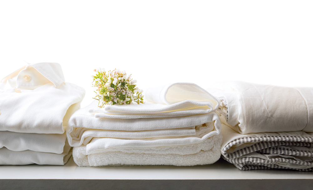 A neatly folded stack of white towels and linens sits on a white surface. A small bouquet of white flowers is placed on top, and a striped fabric is on the right. The background is bright and minimal.