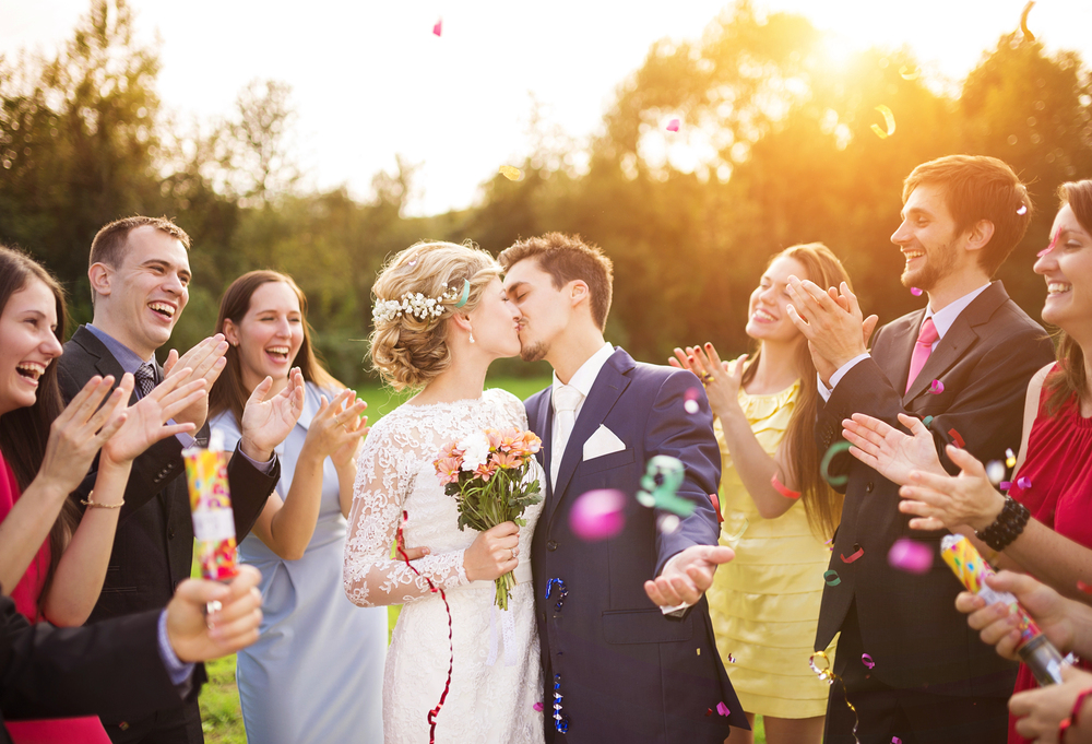 A bride and groom kiss outdoors while smiling friends surround them, clapping and celebrating with confetti. The bride wears a white dress and holds a bouquet, and the group is joyful in the sunlight.