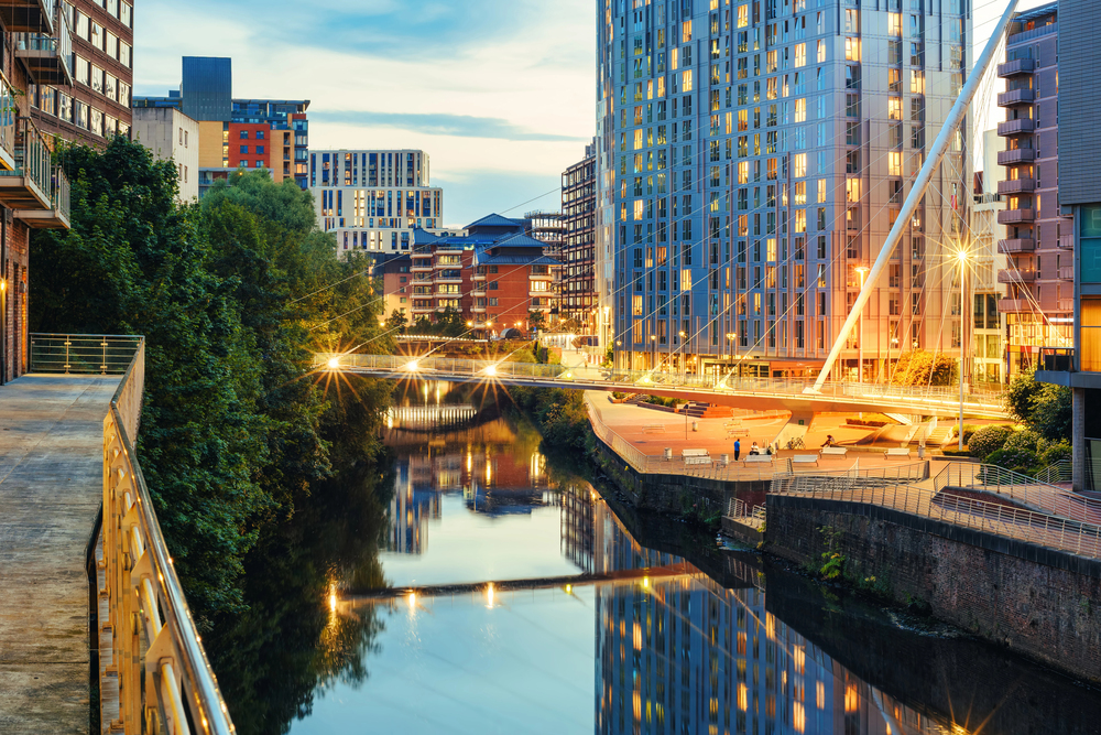A modern cityscape at dusk with tall buildings, a footbridge illuminated by lights, and their reflections visible in a calm river below, surrounded by greenery and urban structures.