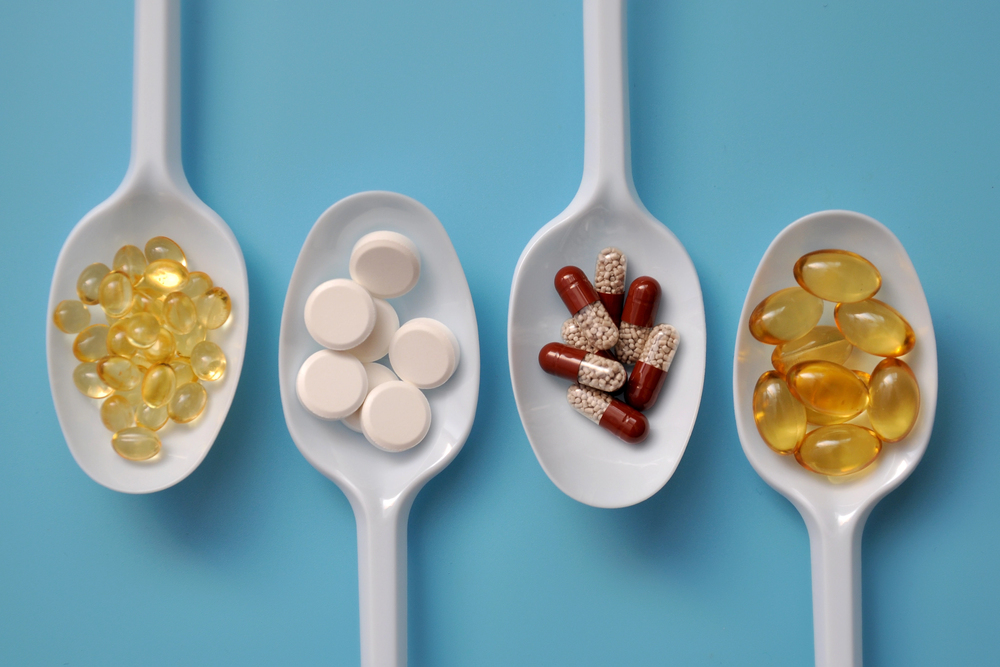 Four white spoons on a blue background, each holding different types of supplements: yellow softgel capsules, round white tablets, red capsules with white pellets, and more yellow softgel capsules.